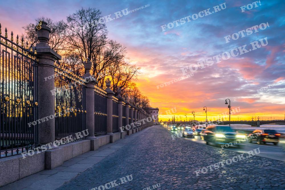 На фотографии изображена набережная реки Невы в Санкт-Петербурге на закате. Видны ограды Летнего сада и проезжающие автомобили. Фотография подходит для использования в туристических материалах, статьях о Санкт-Петербурге и иллюстрациях о городских пейзажах.